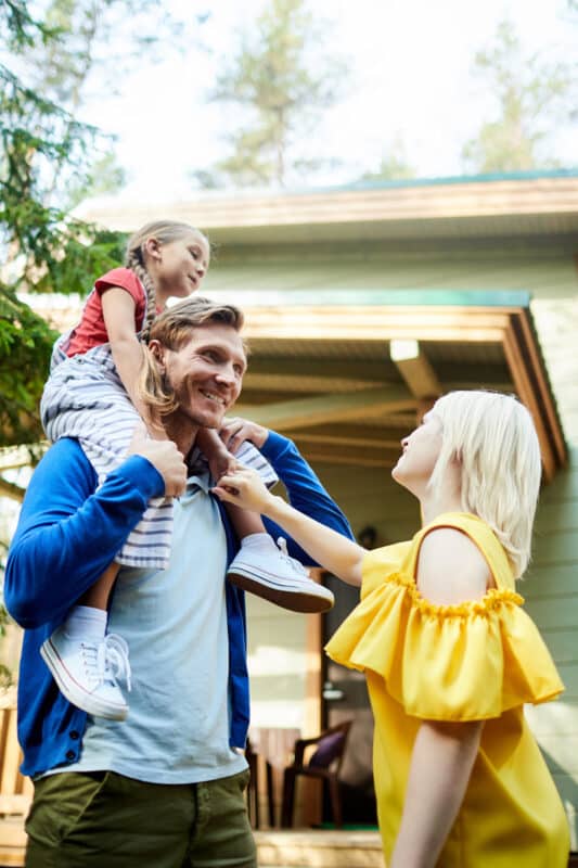 Happy young man with little daughter on his shoulders standing in front of wife while enjoying weekend in the country.