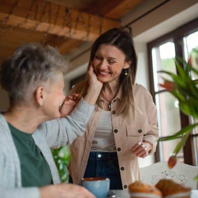 Happy senior mother having tea with adult daughter indoors at home, talking after residential addiction treatment and how to get help for addiction.
